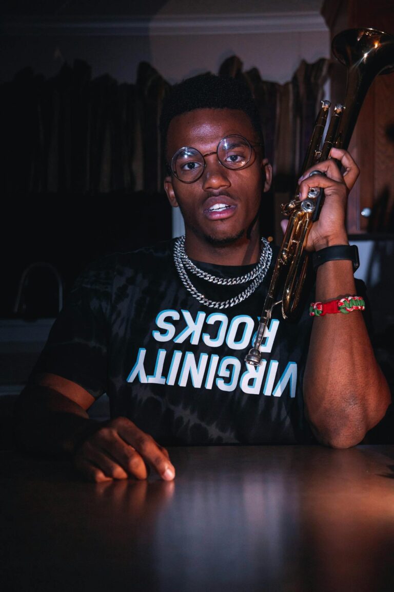 Stylish man holding a trumpet, wearing glasses and vibrant accessories, in an artistic setting.